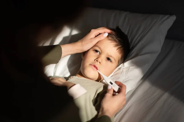 悲しい小さな男の子はベッドの上にあり お母さんは温度計で温度を測定し 暗い寝室のインテリアで額に触れます 健康管理 インフルエンザと風邪の病気 小児期 自宅での治療疾患 — ストック写真