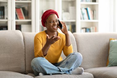 Relaxed candid cheerful stylish young african american woman wearing red turban sitting on couch with legs up at home, talking on phone, gesturing and smiling, looking at copy space