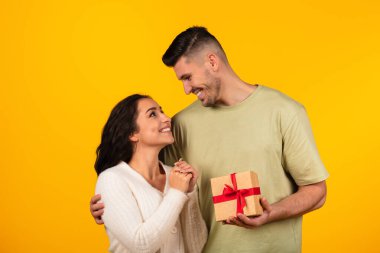 Cheerful young arab guy hugs woman gives box with gift, enjoy romantic moment isolated on yellow background, studio. Congratulations on holiday, anniversary, Valentine day, date, love, relationship