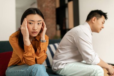 Sad asian wife ignoring upset husband after quarrel, touching head, sitting on sofa in living room interior, selective focus. Relationship problems, crisis, scandal at home