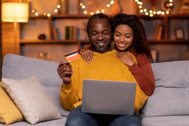 Happy smiling beautiful black lovers shopping from home, sitting on couch in living room decorated with lights, using modern laptop and credit card, copy space. E-commerce concept