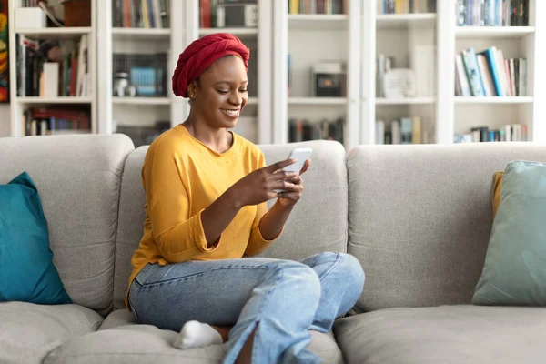 Cheerful relaxed attractive millennial african american woman wearing red turban sitting on couch at home, using modern smartphone, chatting with guys on dating mobile app, shopping online, copy space