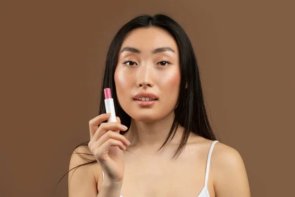 Nude makeup. Portrait of young japanese woman holding pink lipstick in hand and smiling at camera, standing over brown studio background, free space