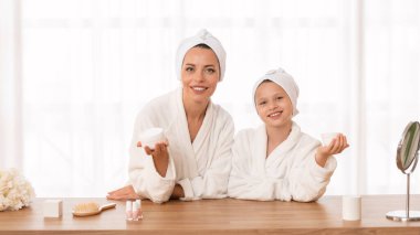 Skin Care Concept. Mother And Little Daughter Wearing Bathrobes Holding Moisturising Cream And Smiling At Camera, Happy Young Mom And Cute Female Child Having Beauty Day At Home, Copy Space