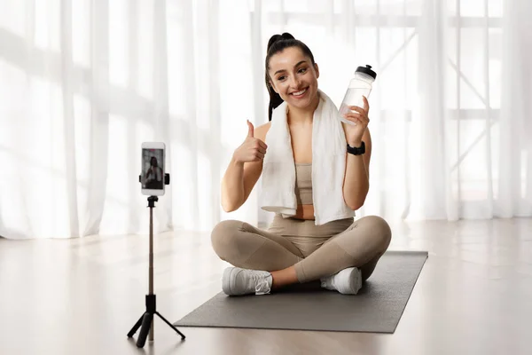 Female fitness coach beautiful good-looking sporty young woman sit on yoga mat with towel on shoulders, showing bottle and thumb up. recommending water drinking, broadcasting from home via smartphone