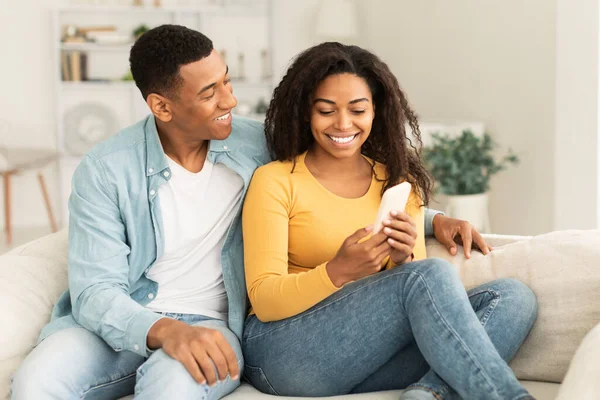 Smiling young african american female show smartphone to male, surfing in internet, chatting in social networks, sitting on sofa in living room interior. New gadget, app recommendation, ad and offer