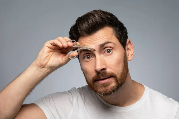 Eyebrow and hair removal of man for skincare. Middle aged bearded man tweezing eyebrows, standing over grey studio background. Cosmetics and self care