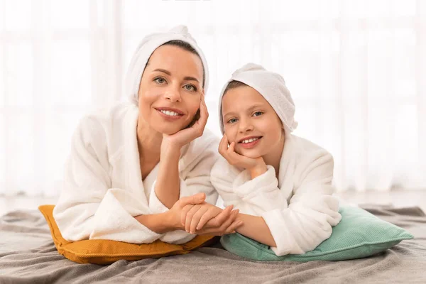 Portrait Of Happy Young Mom And Cute Female Child In Bathrobes Relaxing In Bedroom, Little Daughter And Her Mom Holding Hands And Smiling At Camera, Having Beauty Day At Home, Copy Space