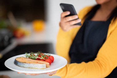 Young african american lady chef in apron makes photo of fresh fish on phone in kitchen interior, blurred, cropped. Professional cooks at home, healthy eat, diet, food blog and app for social networks