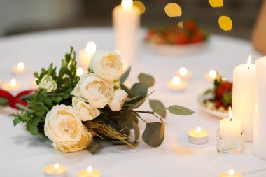 Romantic Dinner. Bouquet of flowers lying on the table, selective focus on bunch of white roses and candles on the desk. Date, Valentine, anniversary concept, blurred background, banner, copy space
