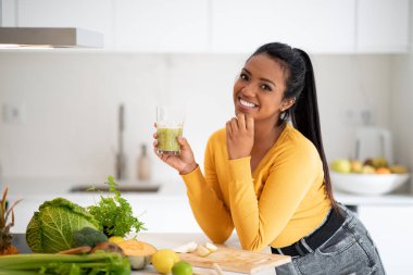 Smiling young african american female vegan at table with organic vegetables hold glass with fresh smoothie in minimalist kitchen interior. Diet, weight loss, cook healthy food at home, body care