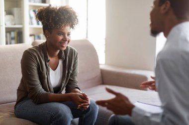 Positive happy young african american lady with bushy hair sitting on couch against black man psychologist, female patient smiling while listening to therapist, successful therapy concept, copy space