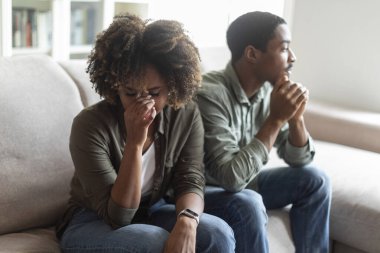 Upset unhappy young black woman with bushy hair crying after quarrel with husband, african american couple experiencing difficulties in relationships, sitting on sofa at home