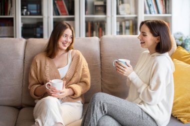 Laughing european millennial ladies in sweaters with coffee cups talk, sit on sofa, enjoy visit, free time in comfort living room interior. Gossip, communication together, relationships and friendship