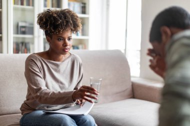 Beautiful young black woman in casual outfit psychologist give male patient glass of water, therapist comforting upset african american guy feeling down during therapy session, copy space