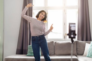 Happy Young Arab Woman Dancing At Camera While Recording Video At Home, Positive Middle Eastern Female Creating Content For Her Social Networks, Using Smartphone On Tripod In Living Room, Copy Space