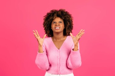 Angry excited upset young black curly female with braces in casual raising her hands and looking up isolated on pink background, studio. Irritation, problems, human emotions and facial expression