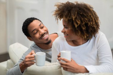 Happy beautiful young black couple in homewear sitting together on couch in living room, drinking coffee, embracing and having conversation, laughing, enjoying lazy weekend at home, copy space
