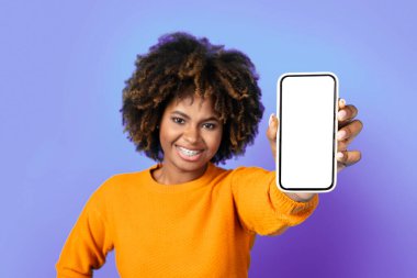 Great online offer. Pretty curly cool young black woman with teeth braces showing brand new cell phone with white empty screen and smiling, isolated on purple studio background, mockup