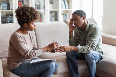 Young african american woman in casual outfit psychotherapist give male patient glass of water, counselor comforting upset african american guy feeling down during therapy session, copy space