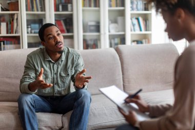 Emotional african american millennial guy in casual sitting on couch at therapist office, sharing feelings and gesturing, young black man attend therapy session with woman psychologist, free space