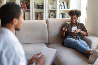 Cheerful positive stylish millennial african american woman reclining on couch at therapist office, talking to black man psychologist, gesturing and smiling, sharing thoughts. Mental health concept