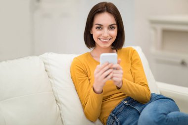 Cute cheerful pretty brunette young woman in casual outfit resting alone on couch at home, using brand new smartphone, checking social media, chatting with friends, smiling at camera, copy space