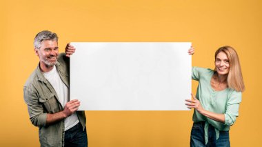 Cheerful middle aged couple holding empty board for advertisement or text and smiling, yellow studio background. Happy man and woman with blank placard for advert