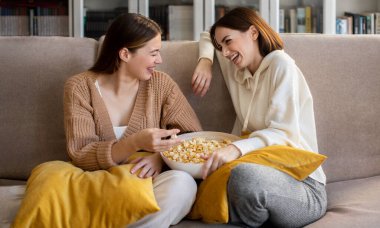 Laughing pretty european millennial girlfriends in sweaters sit on sofa, enjoy popcorn, talk in spare time in comfort living room interior. Relationship, rest together, film at home and communication