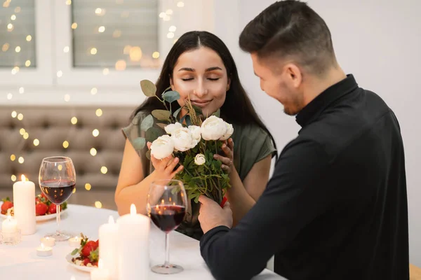 Beautiful millennial couple in love drinking wine, celebrate Valentines day dining at restaurant together, have romantic dinner date with candles sit at table, man giving flowers to woman, copy space