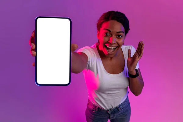 Amazing Offer. Excited Black Woman Showing Blank Smartphone At Camera, Surprised African American Female Demonstrating Empty Cellphone While Standing In Neon Light Over Purple Background, Mockup