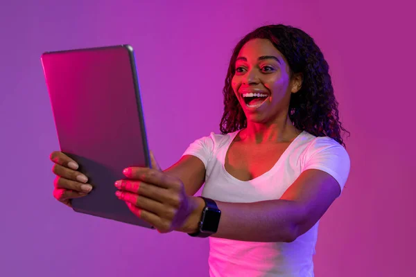 Online Gaming Concept. Surprised black woman looking at digital tablet screen with excitement while standing in neon light over purple background, african american lady emotionally reacting to win