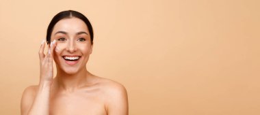 Beauty Banner. Attractive Young Indian Woman Applying Moisturizing Cream On Face, Beautiful Smiling Hindu Lady Rubbing Nourishing Lotion While Standing With Bare Shoulders On Beige Background