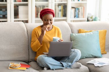 Cheerful young african american lady wearing red turban sitting on couch, drinking tea, eating cookies, watching movie on laptop, female freelancer having break while working from home, copy space
