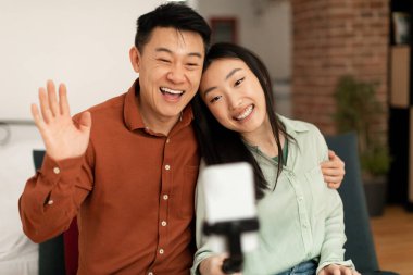 Happy asian spouses making video call via cellphone on tripod, hugging and man waving hands at webcamera, sitting on sofa at home. Husband and wife taking photos on phone