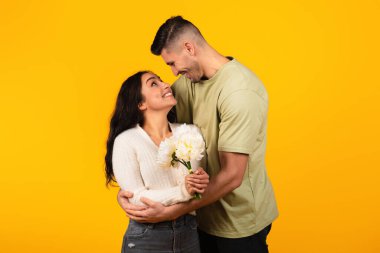 Cheerful young arab guy in casual hugs woman with flowers, enjoy romantic, isolated on yellow background, studio. Love, relationship, congratulations of holiday, Valentine day and anniversary, date