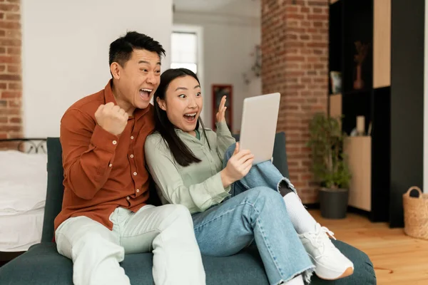Surprised asian spouses using digital tablet, looking at screen with excitement and clenching fists, sitting on sofa at home, free space, living room interior. Couple reading grest news
