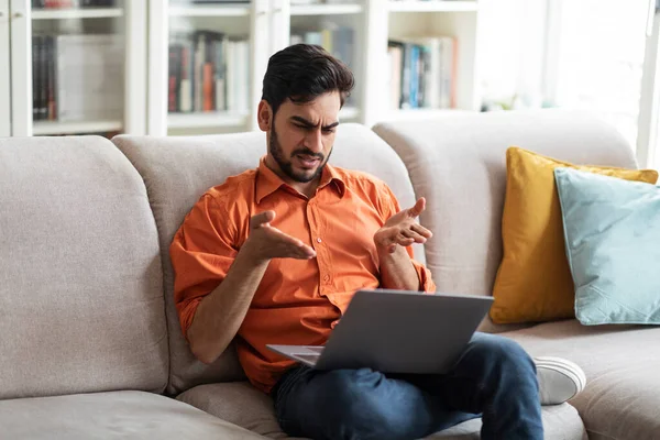 Irritated angry handsome millennial middle eastern man in casual sitting on couch at home, using laptop, looking at computer screen and gesturing, have problems with Internet connection, copy space