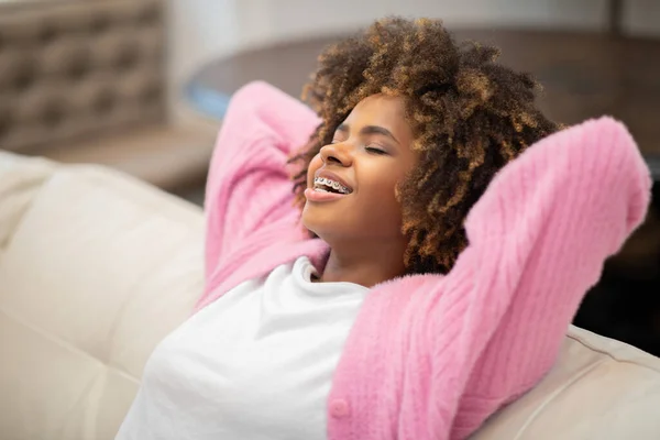 Closeup of relaxed happy pretty young black woman in casual outfit chilling on couch at home, reclining on sofa with closed eyes and arms behind her head, dreaming about wealthy future, copy space