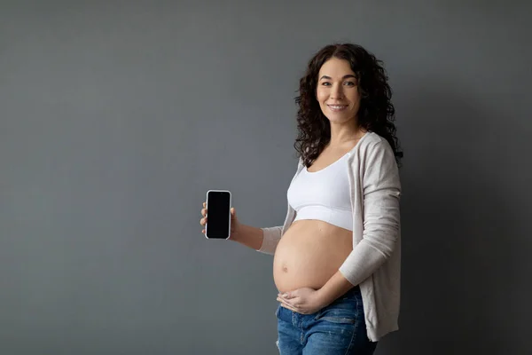 Pregnancy App. Beautiful Young Pregnant Woman Showing Big Blank Smartphone In Hand, Smiling Expectant Female Embracing Belly And Demonstrating Cellphone With Empty Black Screen, Mockup