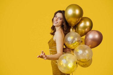 Festive concept. Charming lady in dress with bunch of air balloons having holiday party over yellow studio background, copy space. Cheerful woman having fun on her birthday celebration