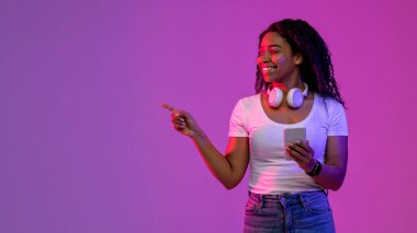 Black Woman With Smartphone In Hand Standing In Neon Light And Pointing Aside At Copy Space Over Purple Studio Background, Young African American Lady Demonstrating Free Place For Advertisement