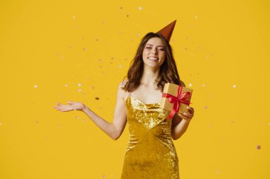 Happy young lady in birthday hat and dress holding gift box, standing over yellow studio background with falling confetti. Pretty woman celebrating holiday, having fun party