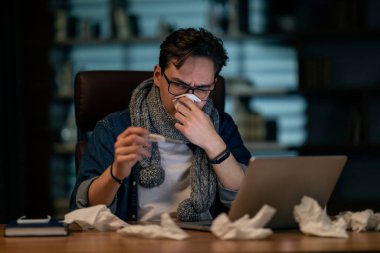 Sick young man in casual wearing eyeglasses and warm scarf around his neck sitting in front of computer at office late at night, sneezing and checking body temperature, holding thermometer, copy space