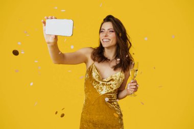 Happy woman in stylish dress taking selfie with glass of champagne, standing over yellow studio background with falling confetti. Positive lady making photo of herself during fun party