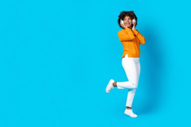 Joyful positive cheerful pretty millennial black lady with bushy hair and teeth braces using wireless headphones and dancing, looking at copy space for ad on blue studio background, panorama