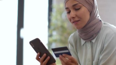 Great mobile banking service. Young happy muslim woman in hijab paying online with smartphone and credit card, entering personal data in app at home, empty space