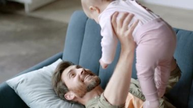 Happy paternity leave. Young playful father playing with his cute baby daughter, lying on sofa and raising her up, tickling kid at home, slow motion, empty space
