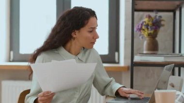 Remote work concept. Young busy middle eastern business lady checking documents and filling online report on laptop, sitting at home office, free space