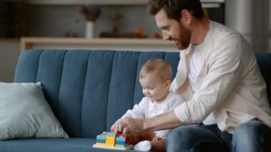 Early development. Caring young father playing with his cute baby daughter at home, searching new wooden toy on sofa, tracking shot, slow motion, empty space